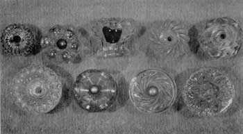 Eight Designs of American Cast Knobs: All are of clear glass. Top row, left to right: Ribbed mushroom design; hollow rosette type and broken knob of same design showing hollow center; swirled mushroom; and design imitating cut glass. Lower row, left to right: Design of fine diamond shapes surrounding deep central cup; hollow crown-shaped rosette; swirl ribs on flat face and conventionalized floral design, also found on Sandwich glass tiebacks. Eight Designs of American Cast Knobs: All are of clear glass. Top row, left to right: Ribbed mushroom design; hollow rosette type and broken knob of same design showing hollow center; swirled mushroom; and design imitating cut glass. Lower row, left to right: Design of fine diamond shapes surrounding deep central cup; hollow crown-shaped rosette; swirl ribs on flat face and conventionalized floral design, also found on Sandwich glass tiebacks.