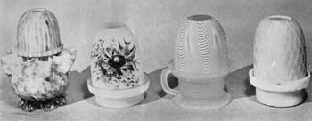 Illustration IV: Four Variations in Night Lights: At the left a specimen attributed to Hobbs, Brockunier Company, Wheeling, West Virginia, of yellow-and-rose flecked glass, a large crimped base and ribbed shade; a lamp of pastel green color and painted decoration depicting a bird and its nest; a rare lamp with handle base in frosted blue glass; and a lamp in pastel yellow mother-of-pearl glass and decorated by diamond quilting on both base and shade. Illustration IV: Four Variations in Night Lights: At the left a specimen attributed to Hobbs, Brockunier Company, Wheeling, West Virginia, of yellow-and-rose flecked glass, a large crimped base and ribbed shade; a lamp of pastel green color and painted decoration depicting a bird and its nest; a rare lamp with handle base in frosted blue glass; and a lamp in pastel yellow mother-of-pearl glass and decorated by diamond quilting on both base and shade.