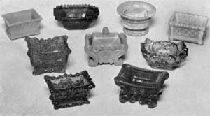 Lacy Sandwich Salt Dishes: Top row, left to right, oblong salt dish of Gothic design in light opaque blue; flaring octagon in light amethyst; circular shape with foot, opaque mottled blue and white; and oblong dish with diaper diamond design, in opalescent. Center row, three of the more elaborate designs in deep amethyst, light opaque blue, and light cobalt blue. Bottom row, salt dish at left in green; and at right, in red. Lacy Sandwich Salt Dishes: Top row, left to right, oblong salt dish of Gothic design in light opaque blue; flaring octagon in light amethyst; circular shape with foot, opaque mottled blue and white; and oblong dish with diaper diamond design, in opalescent. Center row, three of the more elaborate designs in deep amethyst, light opaque blue, and light cobalt blue. Bottom row, salt dish at left in green; and at right, in red.
