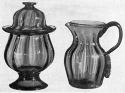 Illustration III: Probably from the Pittsburgh District: The sugar bowl, with cover, has an applied foot and was molded with wide-spaced expanded ribbing. It has a galleried rim, dome cover, flat button finial, and was made of deep sapphire blue glass. The pitcher was molded in the same pattern of blue-colored glass with violet tone. Pieces of this type are believed to have emanated from the Pittsburgh district and, quite possibly, to be a product of the Bakewell, Page & Bakewell Glass Works — period circa 1830-40. Illustration III: Probably from the Pittsburgh District: The sugar bowl, with cover, has an applied foot and was molded with wide-spaced expanded ribbing. It has a galleried rim, dome cover, flat button finial, and was made of deep sapphire blue glass. The pitcher was molded in the same pattern of blue-colored glass with violet tone. Pieces of this type are believed to have emanated from the Pittsburgh district and, quite possibly, to be a product of the Bakewell, Page & Bakewell Glass Works — period circa 1830-40.