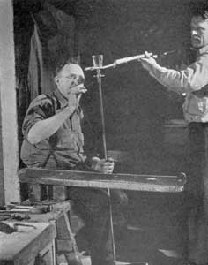 Ready for the Lehr: The goblet is now made and the gaffer is freeing it from the pontil rod as his servitor holds the fork by which the finished piece, still too hot to handle, will be taken to the annealing lehr for the gradual process of cooling. When cold the roughness where the pontil was once attached will be removed at a series of grinding and polishing wheels. Ready for the Lehr: The goblet is now made and the gaffer is freeing it from the pontil rod as his servitor holds the fork by which the finished piece, still too hot to handle, will be taken to the annealing lehr for the gradual process of cooling. When cold the roughness where the pontil was once attached will be removed at a series of grinding and polishing wheels.