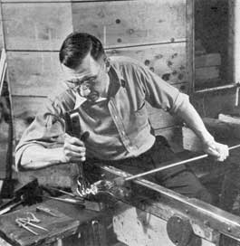 Shaping the Stem: Using a steel fork, as he rolls his blow pipe back and forth on the arms of his bench, the gaffer is giving the goblet stem the desired form. The upper part of the goblet is still attached to the blow pipe and the stem design can be seen. Shaping the Stem: Using a steel fork, as he rolls his blow pipe back and forth on the arms of his bench, the gaffer is giving the goblet stem the desired form. The upper part of the goblet is still attached to the blow pipe and the stem design can be seen.