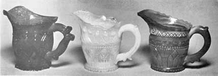 Three Sandwich Cream Pitchers: All are in shades of blue. The right is of streaked opaque glass. The design of that at the left is a combination of the acanthus leaf with diamond-cut shields; that of the one in the center, the Gothic pattern and peacock feather border; and that at the right, the Gothic pattern and the chain border. Three Sandwich Cream Pitchers: All are in shades of blue. The right is of streaked opaque glass. The design of that at the left is a combination of the acanthus leaf with diamond-cut shields; that of the one in the center, the Gothic pattern and peacock feather border; and that at the right, the Gothic pattern and the chain border.