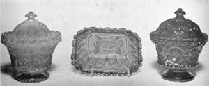 Two Covered Sugar Bowls and a Plate: The sugar bowl at the left is the most elaborate type made at Sandwich. The design combines the acanthus leaf and shield motifs. The upper rim of the bowl part has a delicate foliage design and the foot is scalloped around the bottom. This piece is of cobalt blue. The oblong plate in the center, with design of Gothic motif, is also of cobalt blue. The sugar bowl at the right has a design that combines bull's-eye, a rococo motif, and diamond-cut elements. In color, it is a very deep blue with a purplish cast. Two Covered Sugar Bowls and a Plate: The sugar bowl at the left is the most elaborate type made at Sandwich. The design combines the acanthus leaf and shield motifs. The upper rim of the bowl part has a delicate foliage design and the foot is scalloped around the bottom. This piece is of cobalt blue. The oblong plate in the center, with design of Gothic motif, is also of cobalt blue. The sugar bowl at the right has a design that combines bull's-eye, a rococo motif, and diamond-cut elements. In color, it is a very deep blue with a purplish cast.