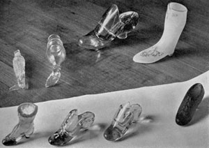 Illustration V: A Variety of Slippers, Boots and Shoes: In upper row are two perfume bottles, an unusual slipper, and an opaque boot decorated with gold, probably of English workmanship. Lower row, a group of thimble holders. The two at the center were made as souvenirs that were sold at the Buffalo Pan-American Exposition. Illustration VI: More Boots and Slippers: Of the two high boots in the center, that at left has a view of Hamburg at the top of the leg. The slippers at the right are rarities as are the smaller frosted boots at the extreme left.