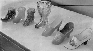 Illustration IV: Shoes and Slippers in Glass: At the left are three shoes in colored case glass with applied leaves in small festoons. The three slippers at the right are of opaque glass, two of which have hand painted decoration. Illustration IV: Shoes and Slippers in Glass: At the left are three shoes in colored case glass with applied leaves in small festoons. The three slippers at the right are of opaque glass, two of which have hand painted decoration.