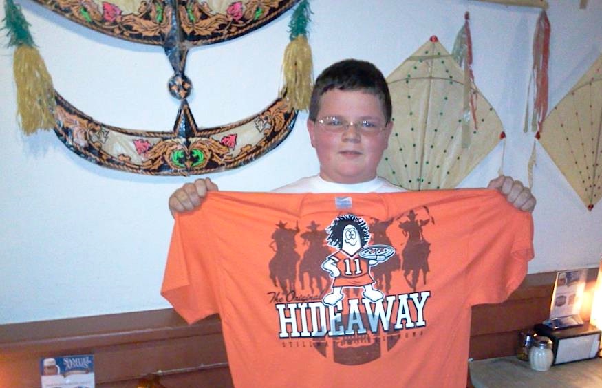 hideaway Nash, celebrating his 14th birthday in the Stillwater restaurant in 2011, holds up a Hideaway Pizza T-shirt with an Oklahoma State Cowboys theme. The wall behind him features a "wau bulan" moon kite from Malaysia and two "pakpao" female kites from Thailand. Via the original Hideaway Pizza Facebook page.