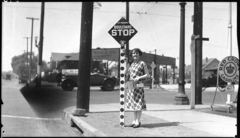 A woman poses with a newly installed stop sign in Los Angeles in 1925, built to the specifications recommended at the first National Conference on Street Safety. Via USC Libraries.
