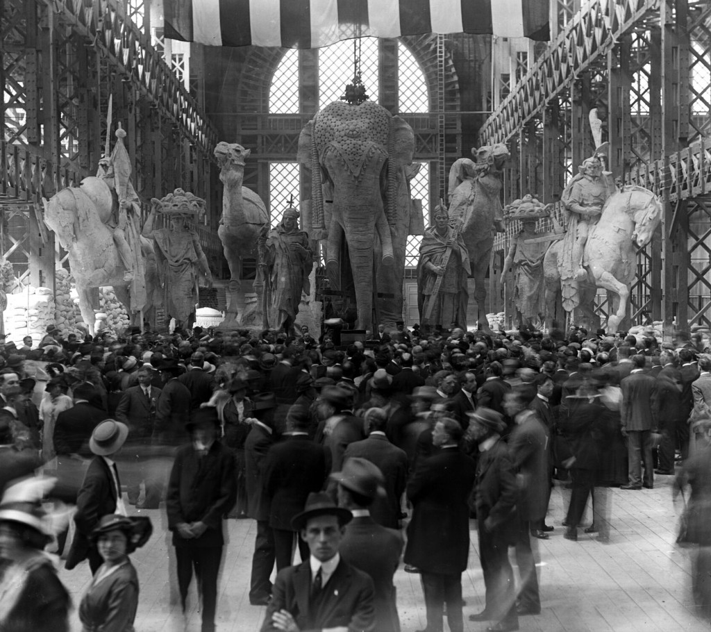 In one of the unfinished palaced, the sculpture group called "Nations of the East" awaits installation over the Arch of the Rising Sun behind a crowd previewing the fairgrounds.