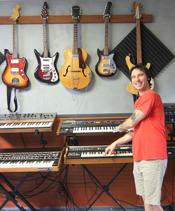 Lance Hill, goofing around with a pair or Roland synthesizers. The vintage guitars on the wall behind him are also used by artists who come to his studio to record.